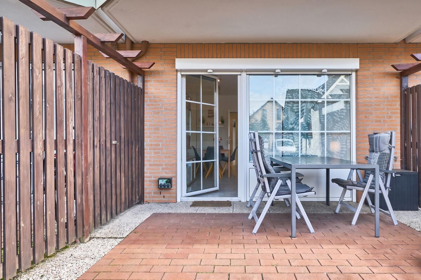 Terrasse mit Tisch und Stühlen, Glasfront zur Wohnung, Holzgeländer und Ziegelsteinmauer.