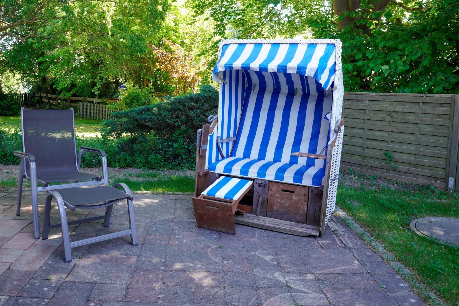 Ein blau-weißer Strandkorb und ein Sitzmöbel auf einem gepflasterten Hof mit Grünflächen und Bäumen.