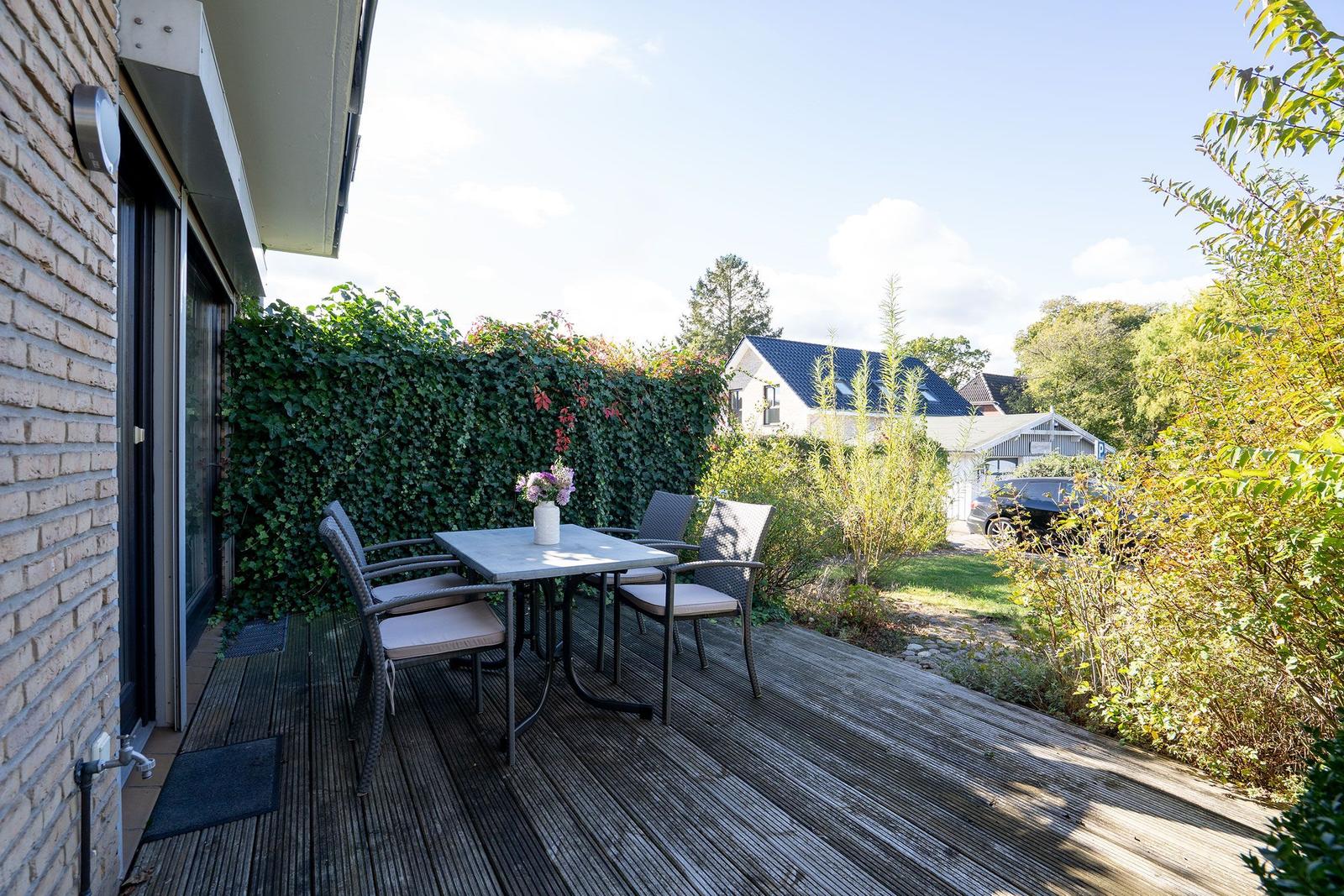 Terrasse mit Tisch und vier Stühlen, umgeben von grüner Hecke und Pflanzen.