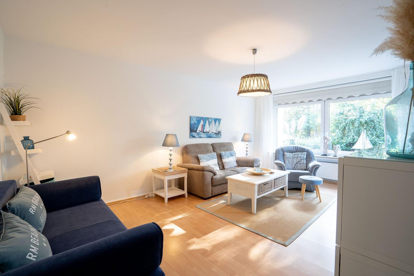 Wohnzimmer mit Sofa, Sessel, Tisch und Fenster. Holzboden und Wandbild mit Segelbooten.