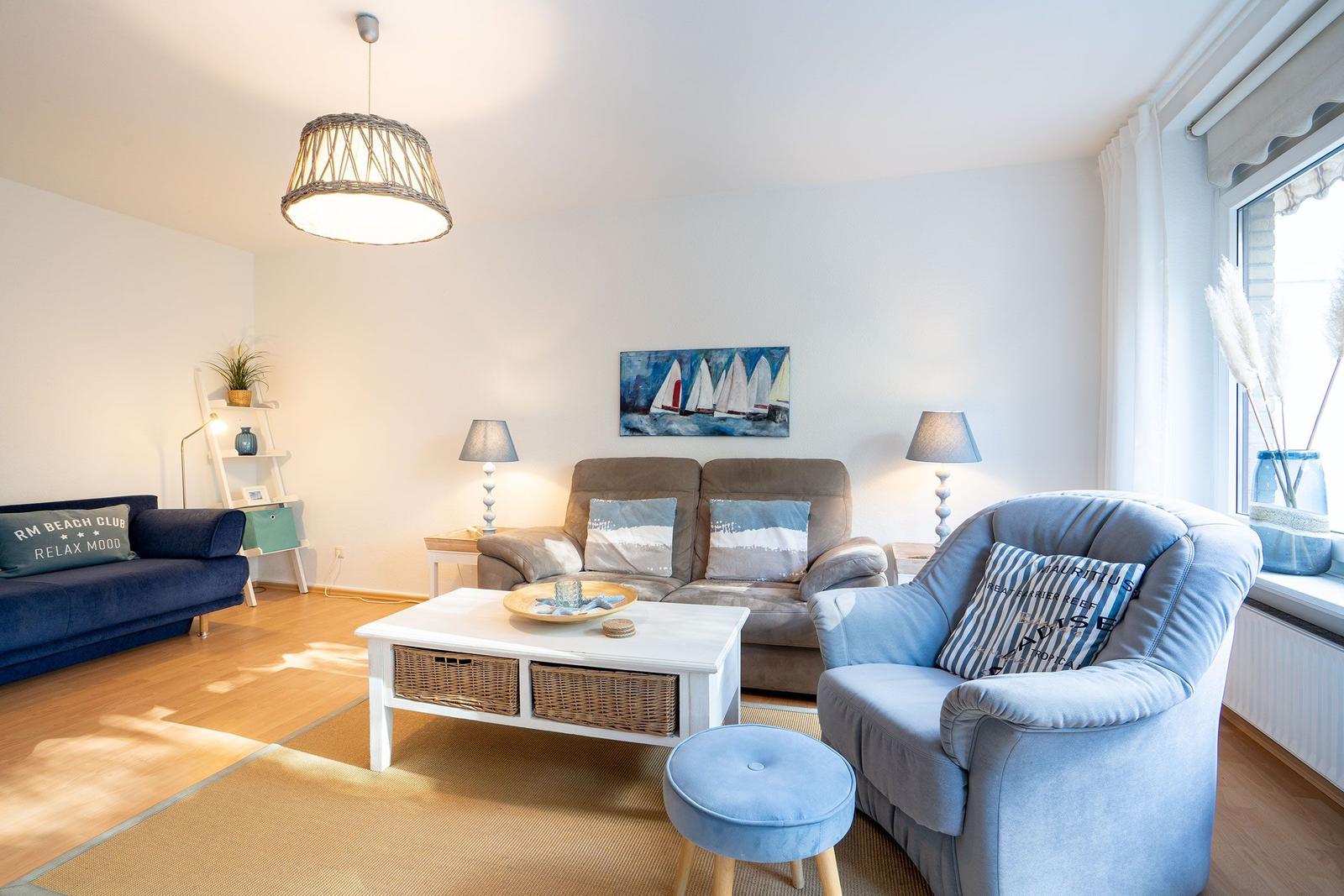 Wohnzimmer mit Sofa, Sessel und Tisch. Wandbild mit Segelbooten. Fenster mit Vorhängen.