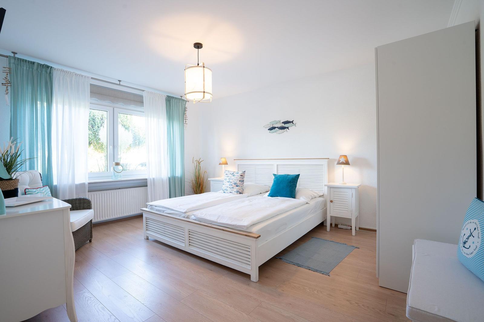 Schlafzimmer mit Doppelbett, Fenster, Nachttischen und Gardinen.