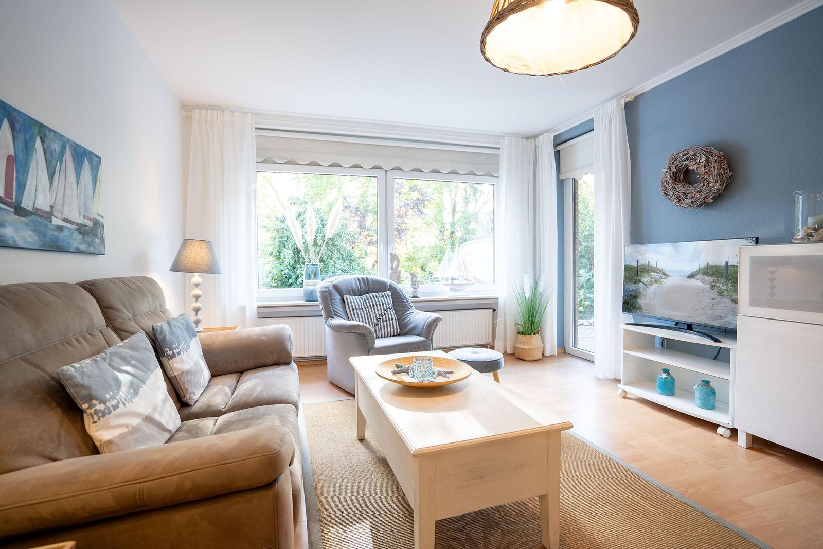 Wohnzimmer mit Sofa, Sessel und Fernseher. Fenster mit Blick auf Grün.