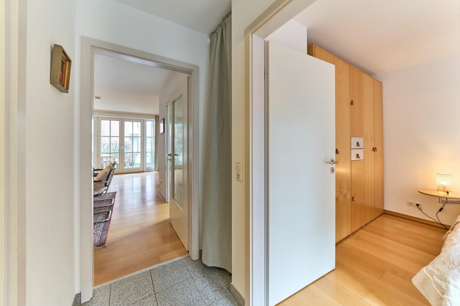 Flur mit Blick in Schlafzimmer und Wohnbereich mit Holzboden und Gardinen.