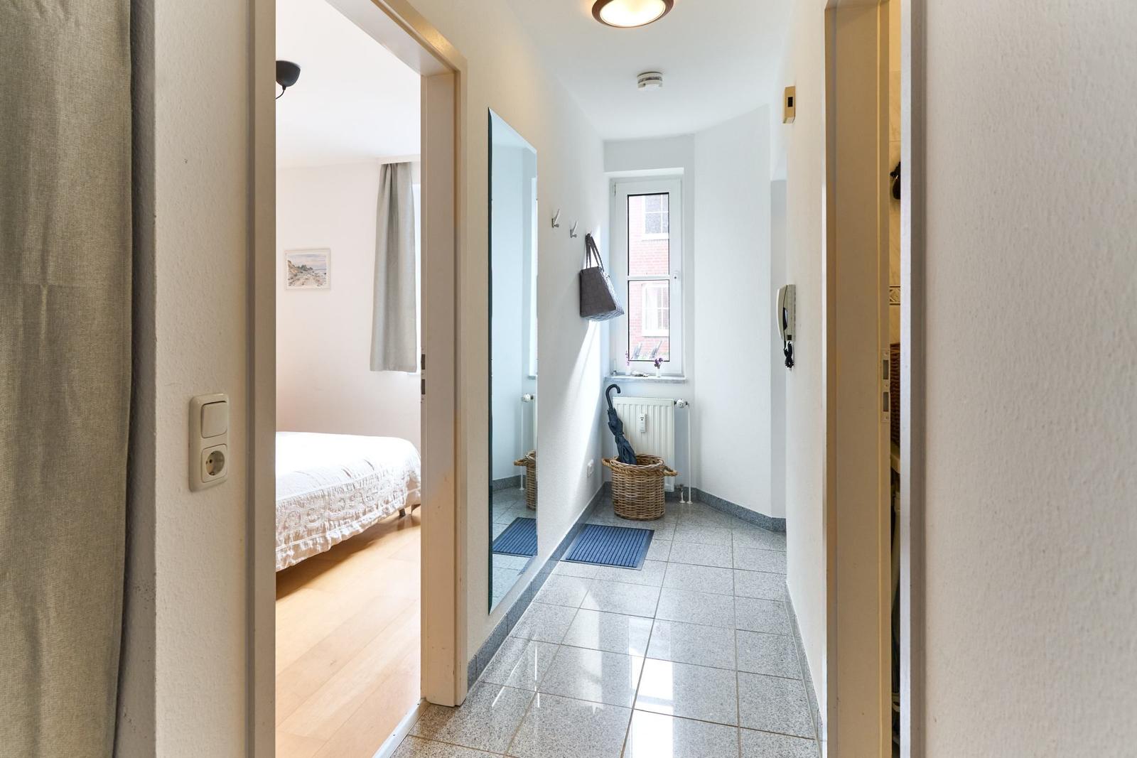 Flur mit Spiegel, Fenster und Wäschekorb. Im Hintergrund Schlafzimmer mit Bett.