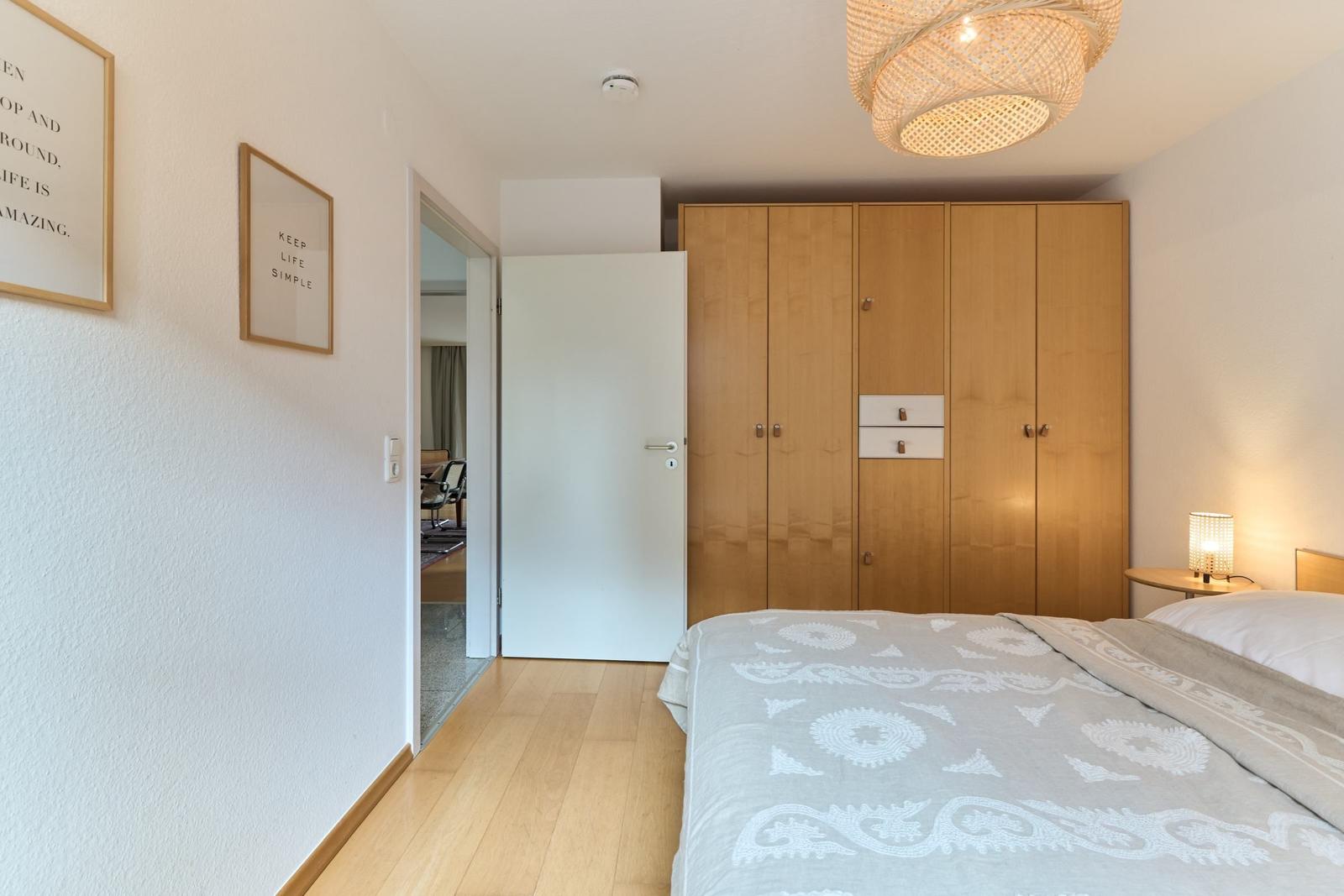 Schlafzimmer mit Bett, Holzschrank und Deckenleuchte. An der Wand hängen zwei Bilder.