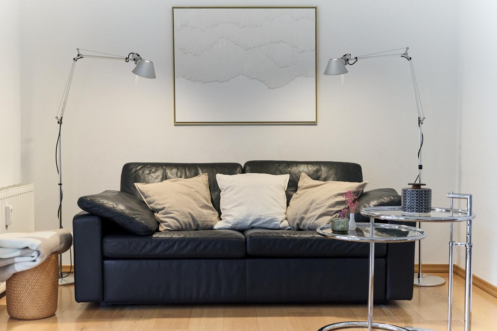 Schwarzes Ledersofa mit Kissen, zwei Stehlampen und Glas-Tisch.