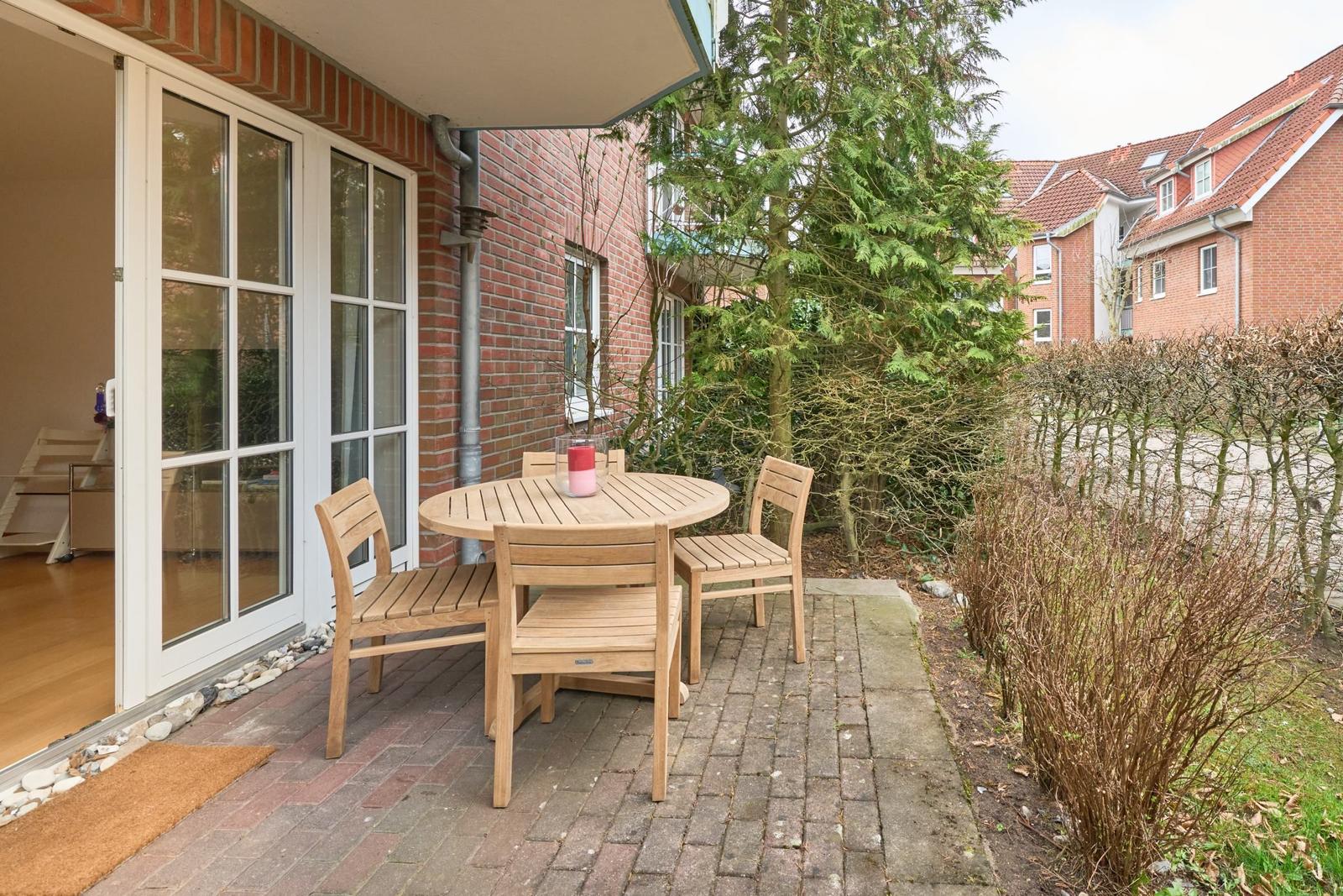 Terrasse mit Holztisch und Stühlen, angrenzend an ein Haus mit Glasfenstern.