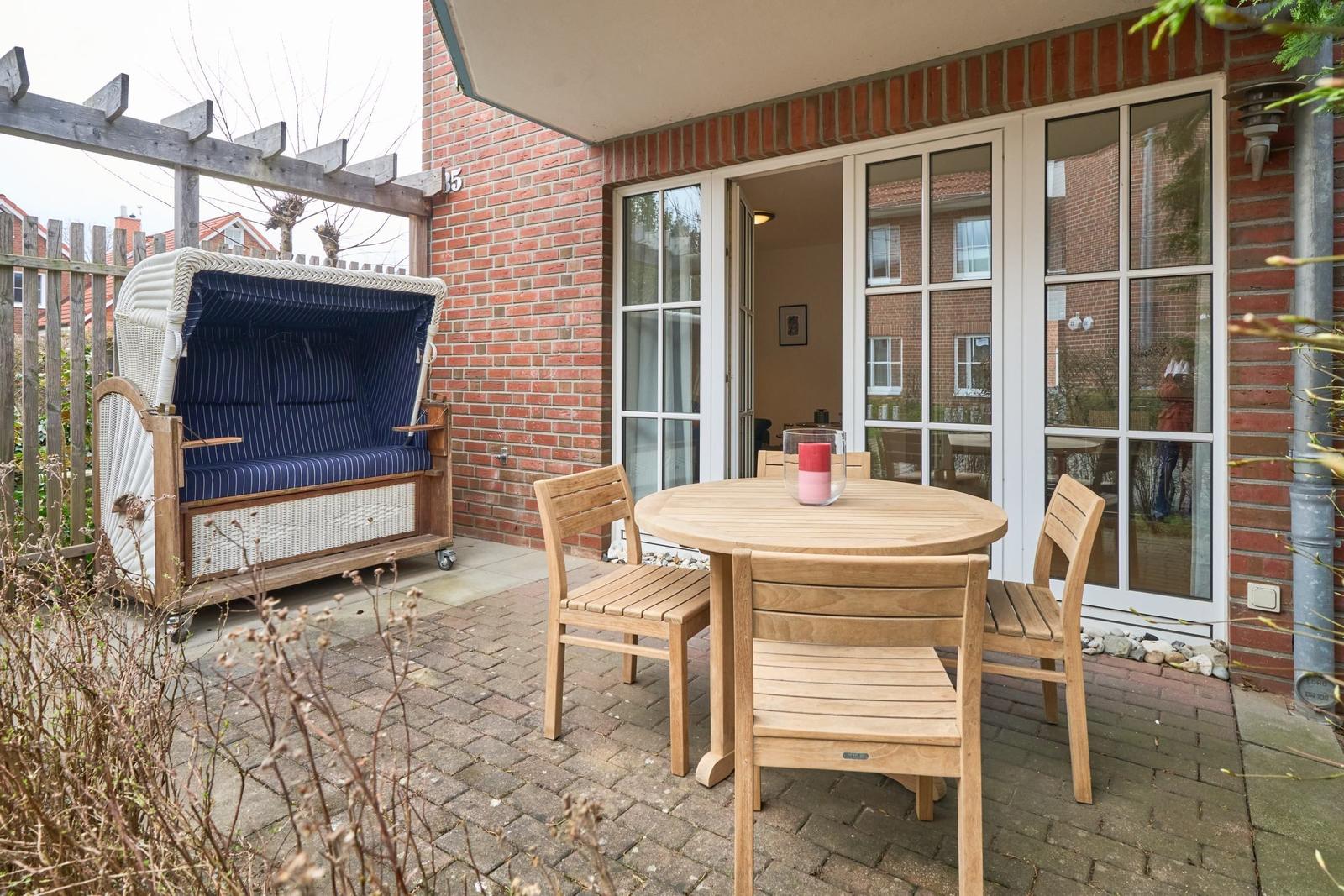 Terrasse mit Holztisch, Stühlen und Strandkorb. Hintergrund: Haus mit Glasfront.
