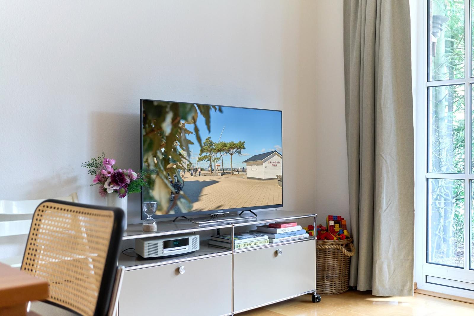 TV auf Konsole mit Blumen und Spielzeugkorb. Fenster mit Vorhang. Fernseher zeigt Strand.