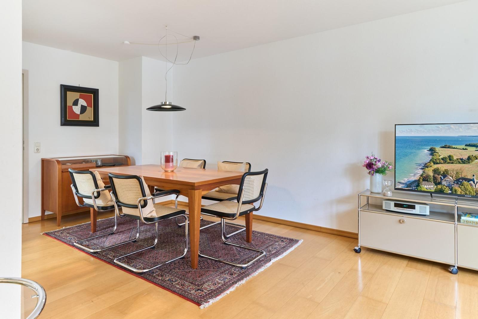 Esszimmer mit Holztisch, Stühlen und TV. Teppich und Wandbild sichtbar.