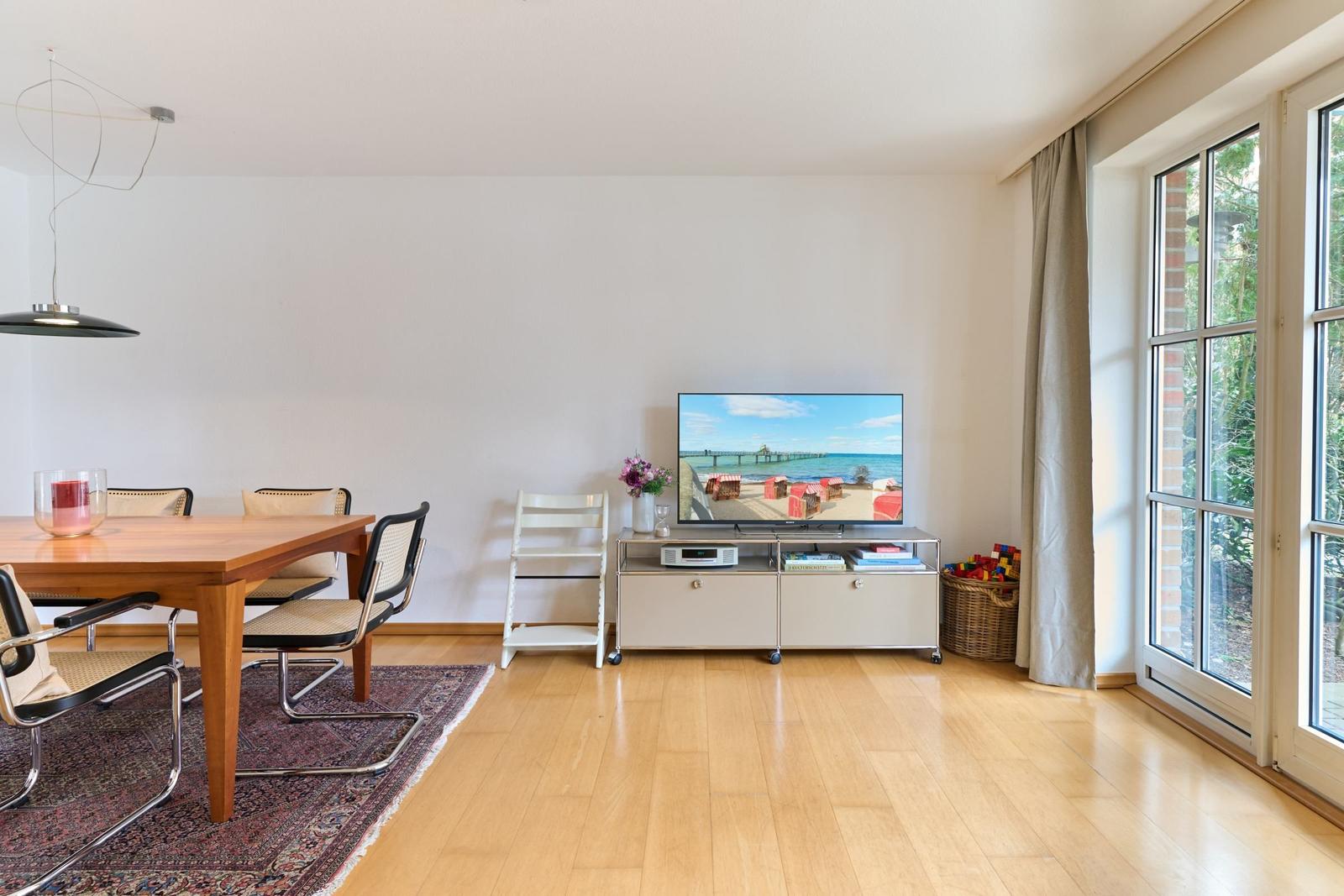 Wohn- und Essbereich mit TV, Holzboden und Fenster. Esstisch mit Stühlen und Teppich.