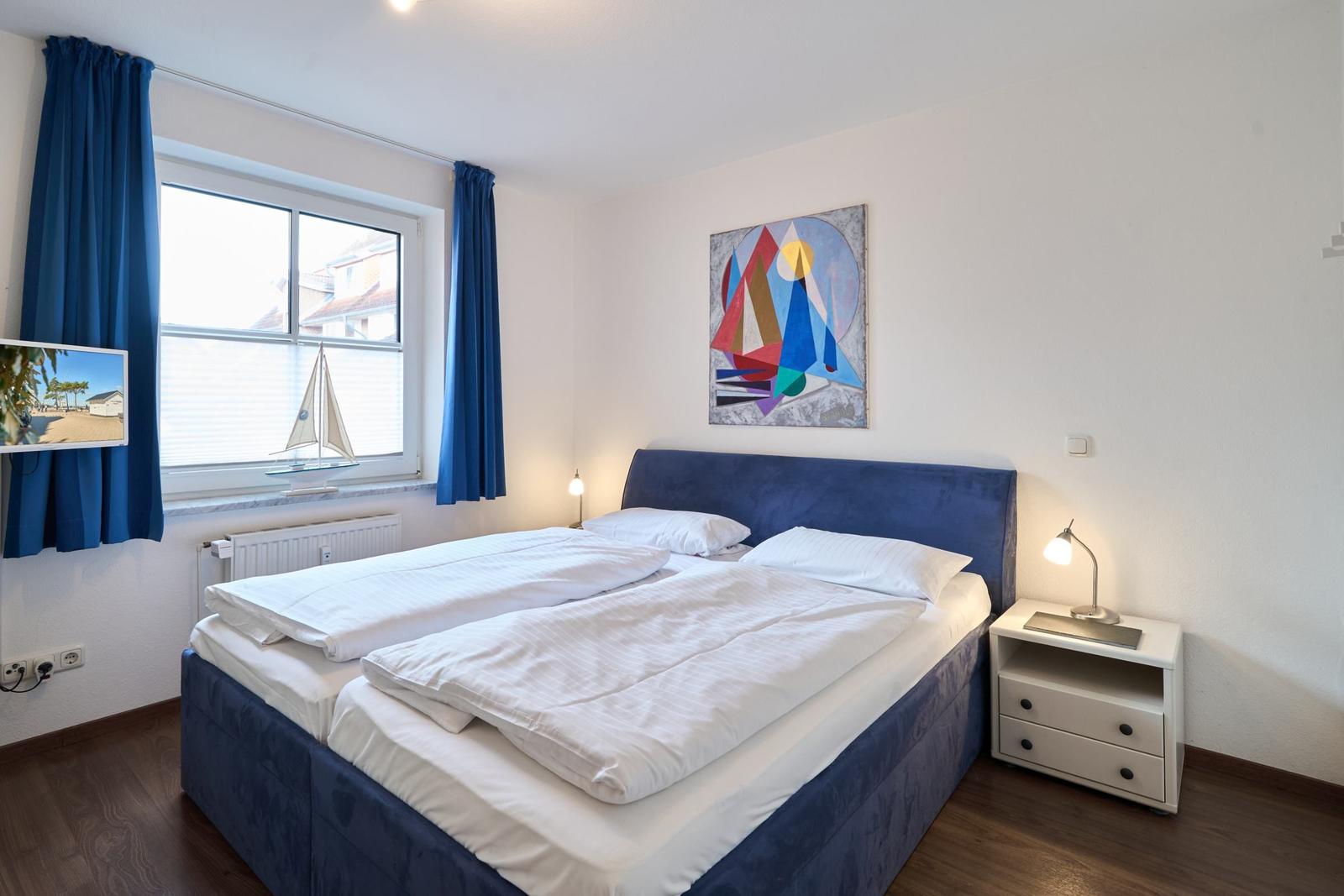 Schlafzimmer mit Doppelbett, blauen Vorhängen und Wandbild. Fenster mit Segelbootmodell.