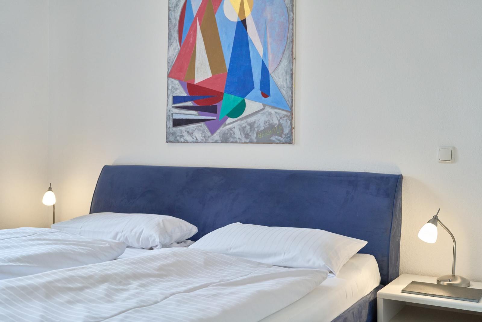 Doppelbett mit blauem Kopfteil, weiße Bettwäsche und abstraktes Wandbild.