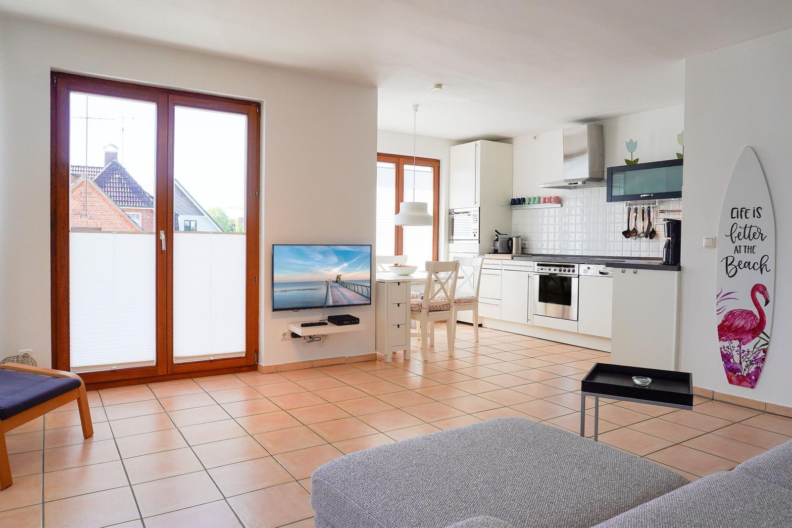 Wohnküchenbereich mit TV, Esstisch und Küchenzeile. Terrassentür und Surfbrett an der Wand.