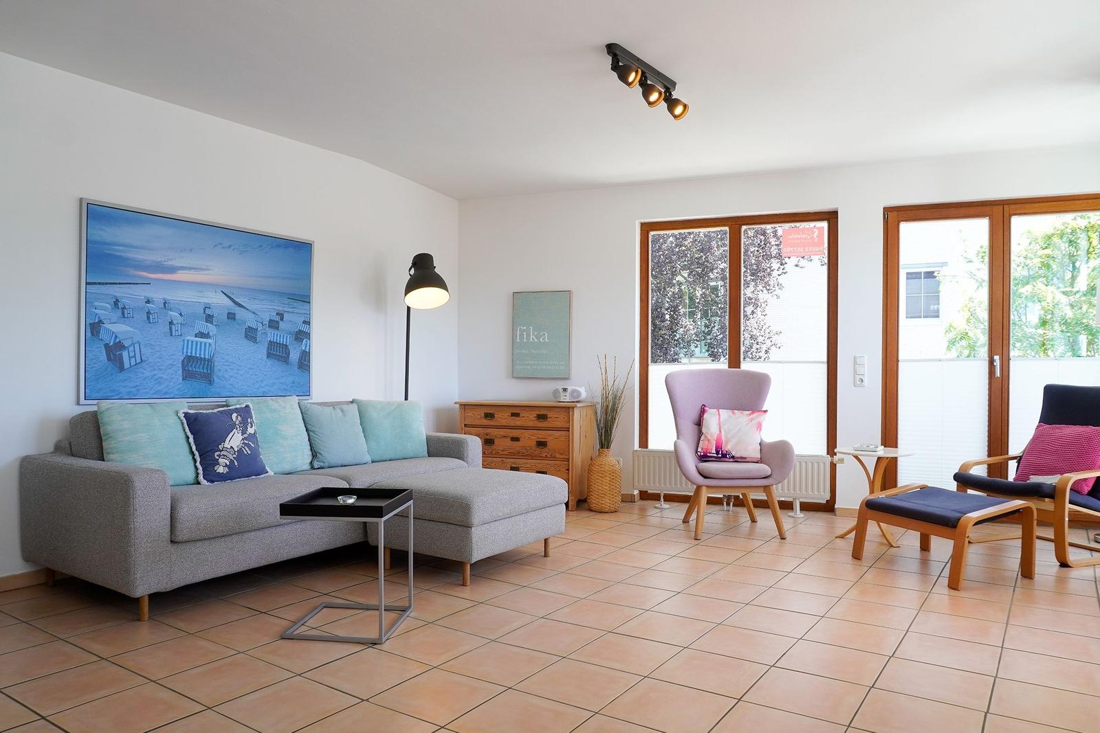 Wohnzimmer mit Sofa, Sessel und großen Fenstern. Wandbild zeigt Strand.