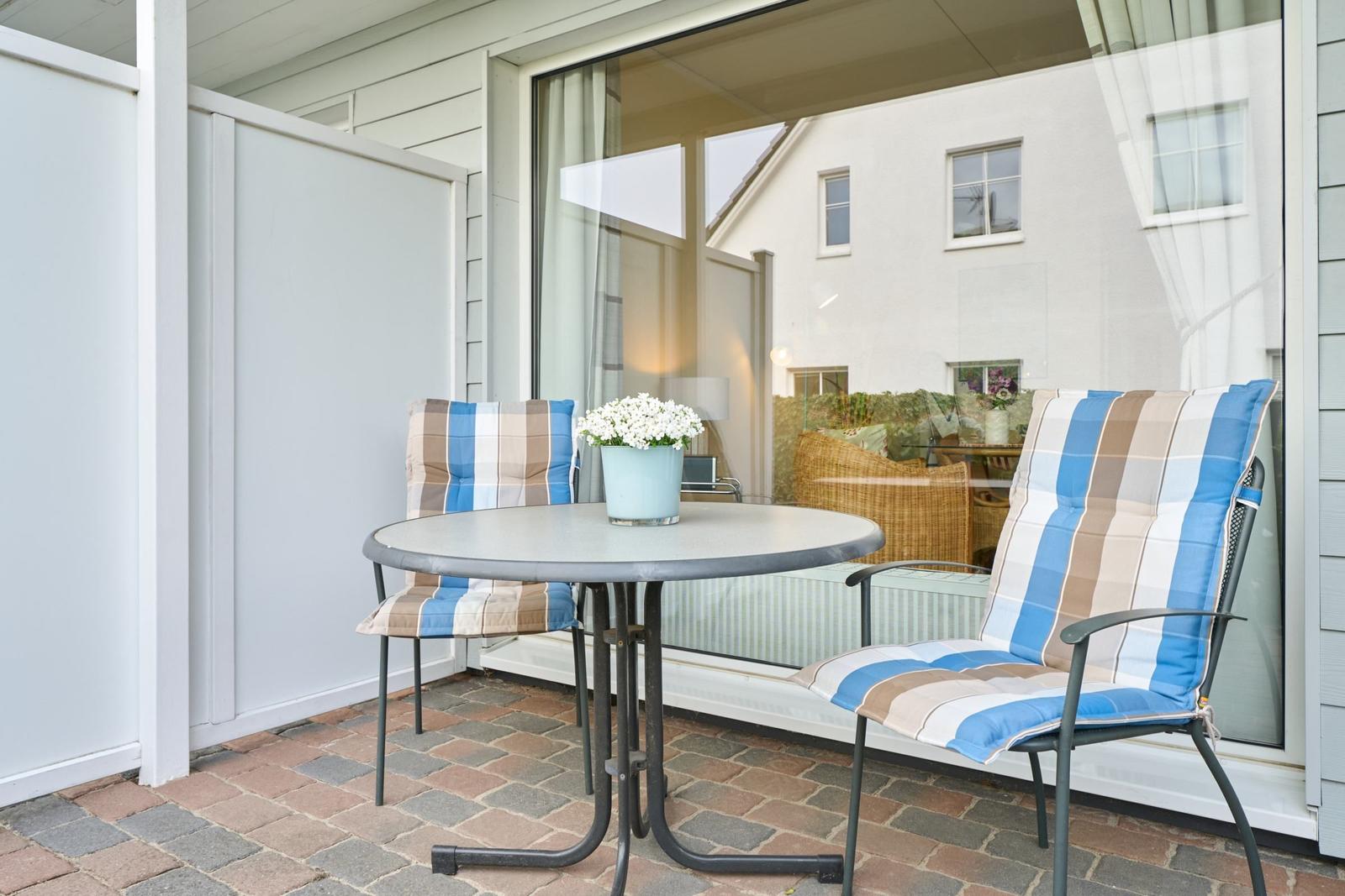 Terrasse mit runden Tisch und zwei Stühlen mit karierten Kissen. Im Hintergrund Glasfront und Haus.