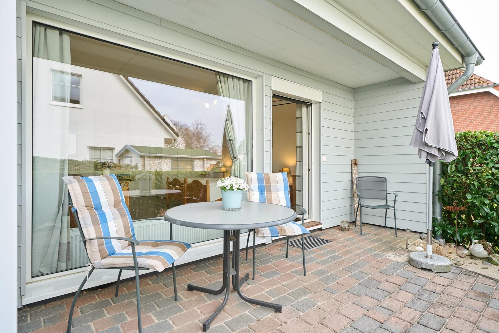 Terrasse mit Tisch, zwei Stühlen und geschlossenem Sonnenschutz. Durchgehende Glasfront zum Innenraum.