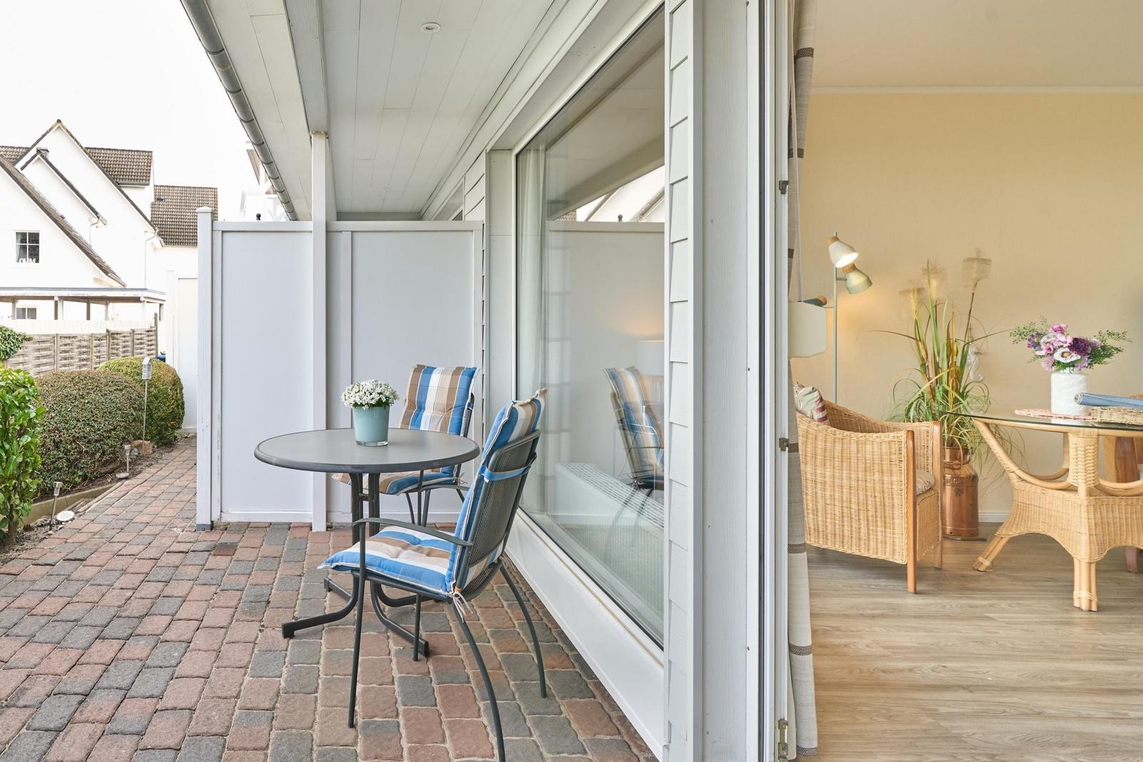 Terrasse mit Tisch und Stühlen. Durch Glastür ist Wohnbereich mit Rattanmöbeln sichtbar.