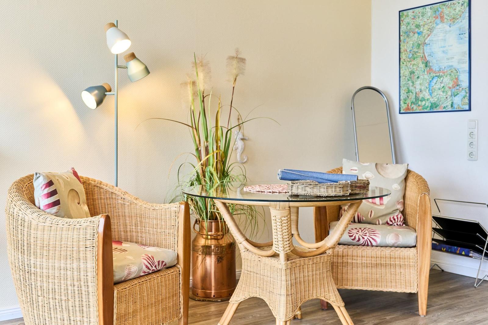 Zwei geflochtene Sessel um einen runden Tisch mit Glasplatte. Hintergrund: Wandlampe und Pflanze.