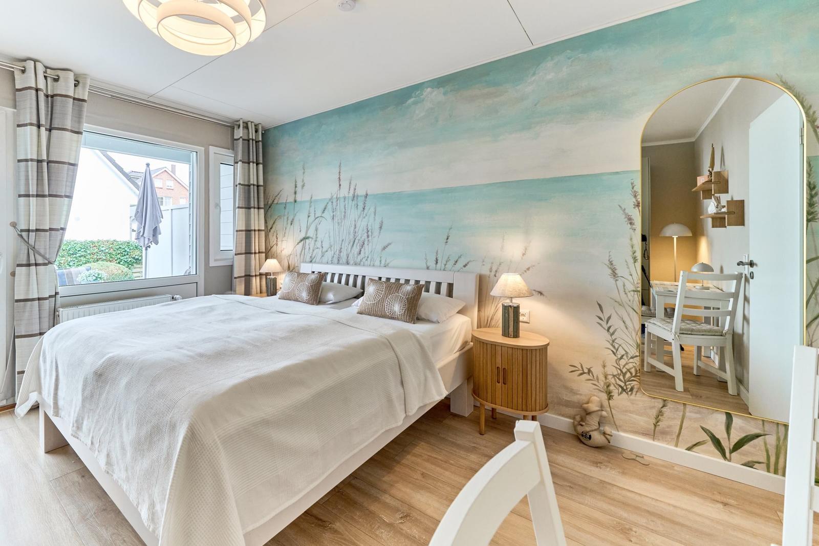 Schlafzimmer mit Bett, Wandmalerei und Spiegel. Fenster mit Vorhängen und Nachbarhaus sichtbar.