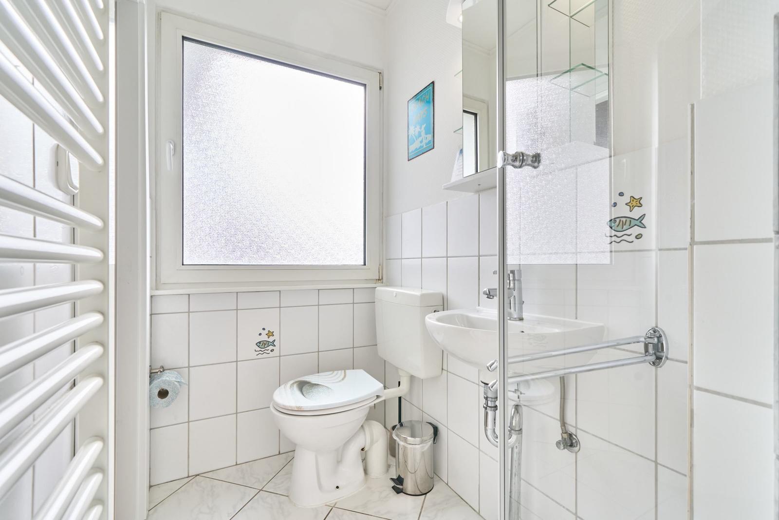 Badezimmer mit Toilette, Waschbecken und Dusche. Fenster mit Milchglas und weiße Fliesen.