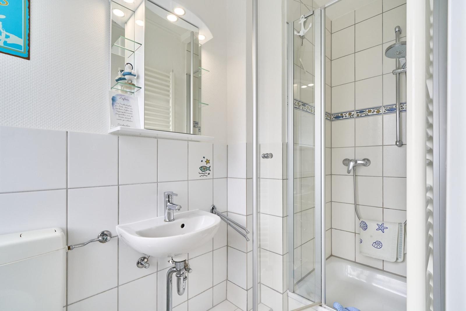 Badezimmer mit Dusche, Waschbecken und Spiegel. Wandfliesen und Handtuchhalter sichtbar.