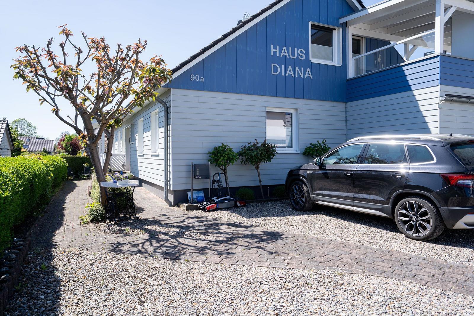 Haus Diana mit blau-weißem Holzschuppen, gepflasterter Hof und parkendem Auto.