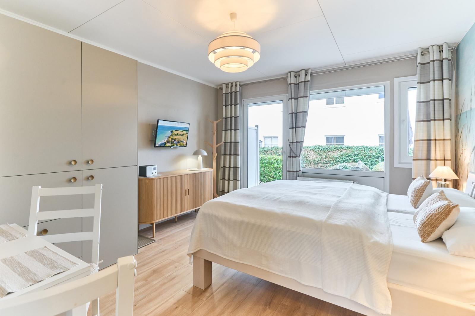 Schlafzimmer mit Bett, TV, Schrank und Fenster mit Blick auf Grünfläche.