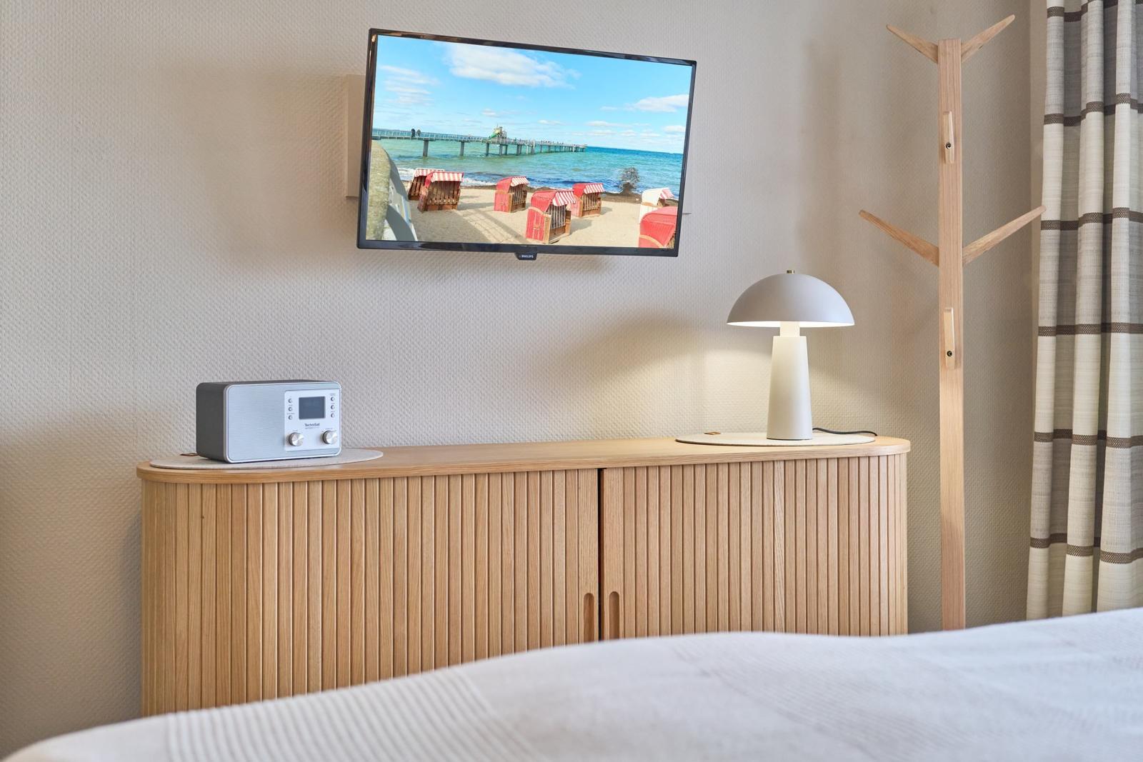 TV an der Wand, daneben Lampe und Radio auf Holzschrank