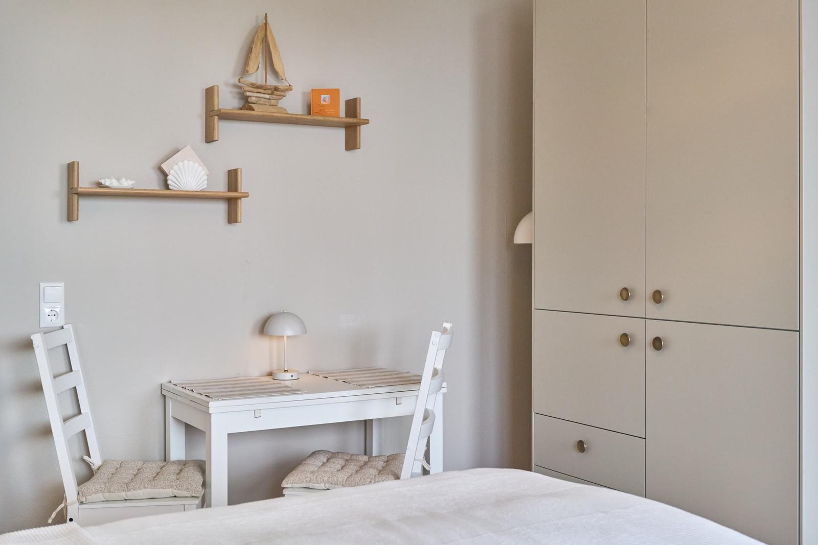 Ein Schreibtisch mit zwei Stühlen und einer Lampe. An der Wand hängen zwei Holzregale mit Dekoration.