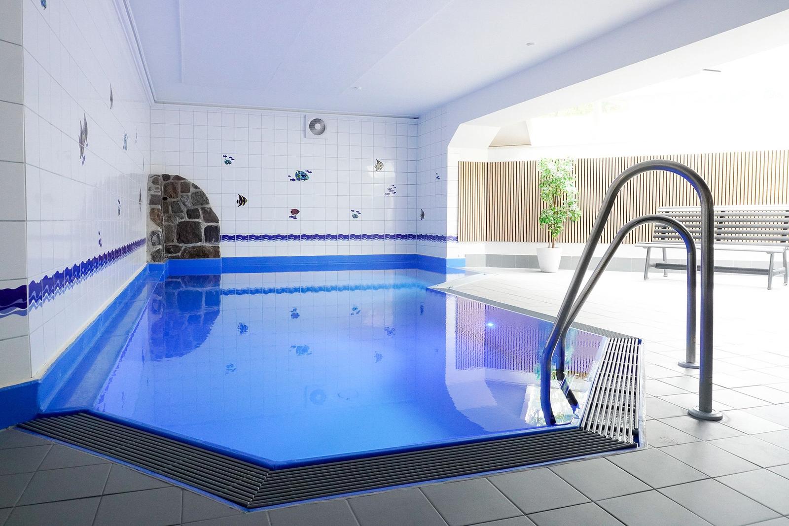 Innere Schwimmhalle mit blauem Wasser, Wandfliesen und Steintreppe.