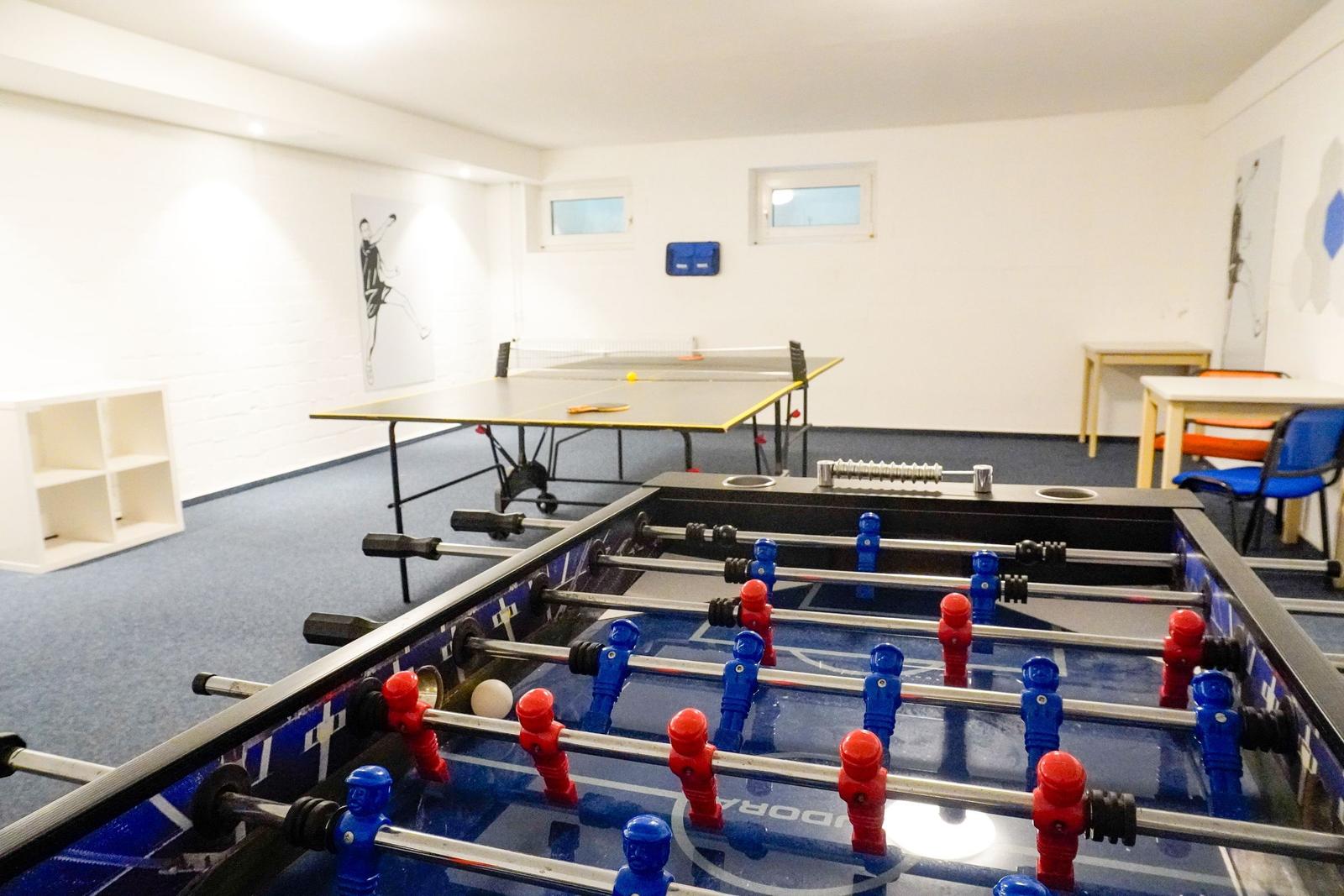 Raum mit Tischfußball und Tischtennisplatte. Blauer Teppich und weiße Wände.