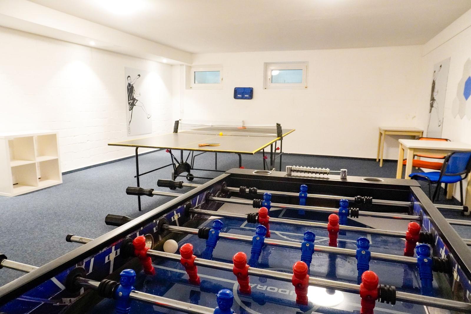 Raum mit Tischtennisplatte und Fussballtisch. Weiße Wände, blauer Teppich und Kunstwerke an den Wänden.