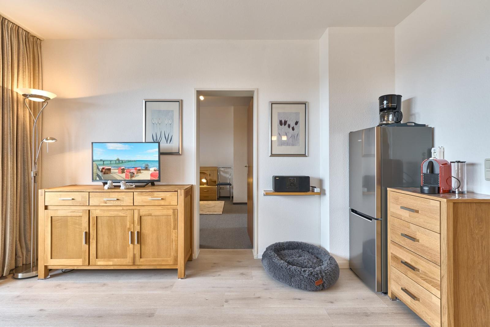 Wohnzimmer mit Fernseher, Kühlschrank und Schlafzimmer im Hintergrund.