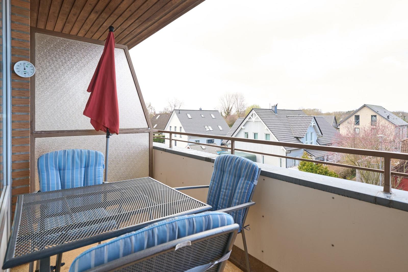 Terrasse mit Tisch, drei Stühlen und rotem Sonnenschirm. Blick auf Wohnhäuser und Bäume.