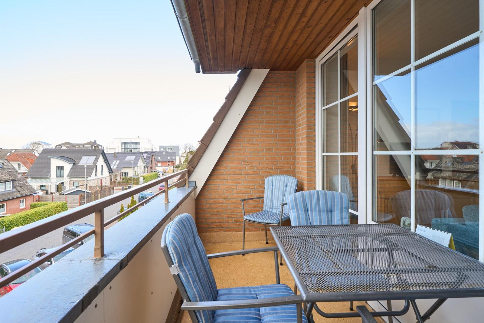 Balkon mit Tisch und drei Stühlen, Blick auf Wohngebiet mit Dächern und Bäumen.
