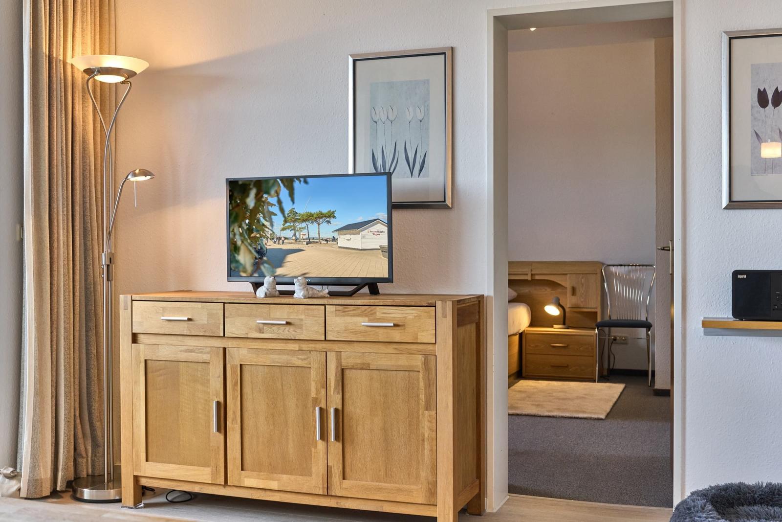 Ein Wohnbereich mit Holzschrank, Fernseher und Stehlampe. Im Hintergrund ist ein Schlafzimmer sichtbar.