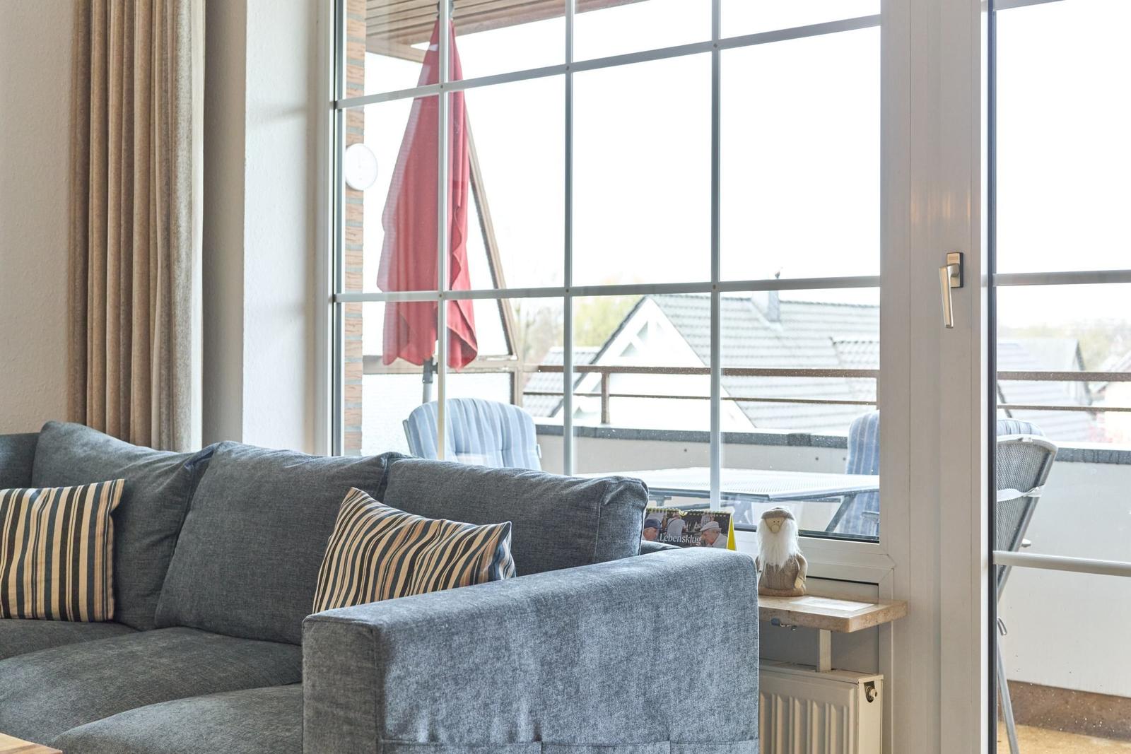 Grauer Sofa mit Kissen vor großem Fenster zum Balkon mit Tisch und Stühlen.