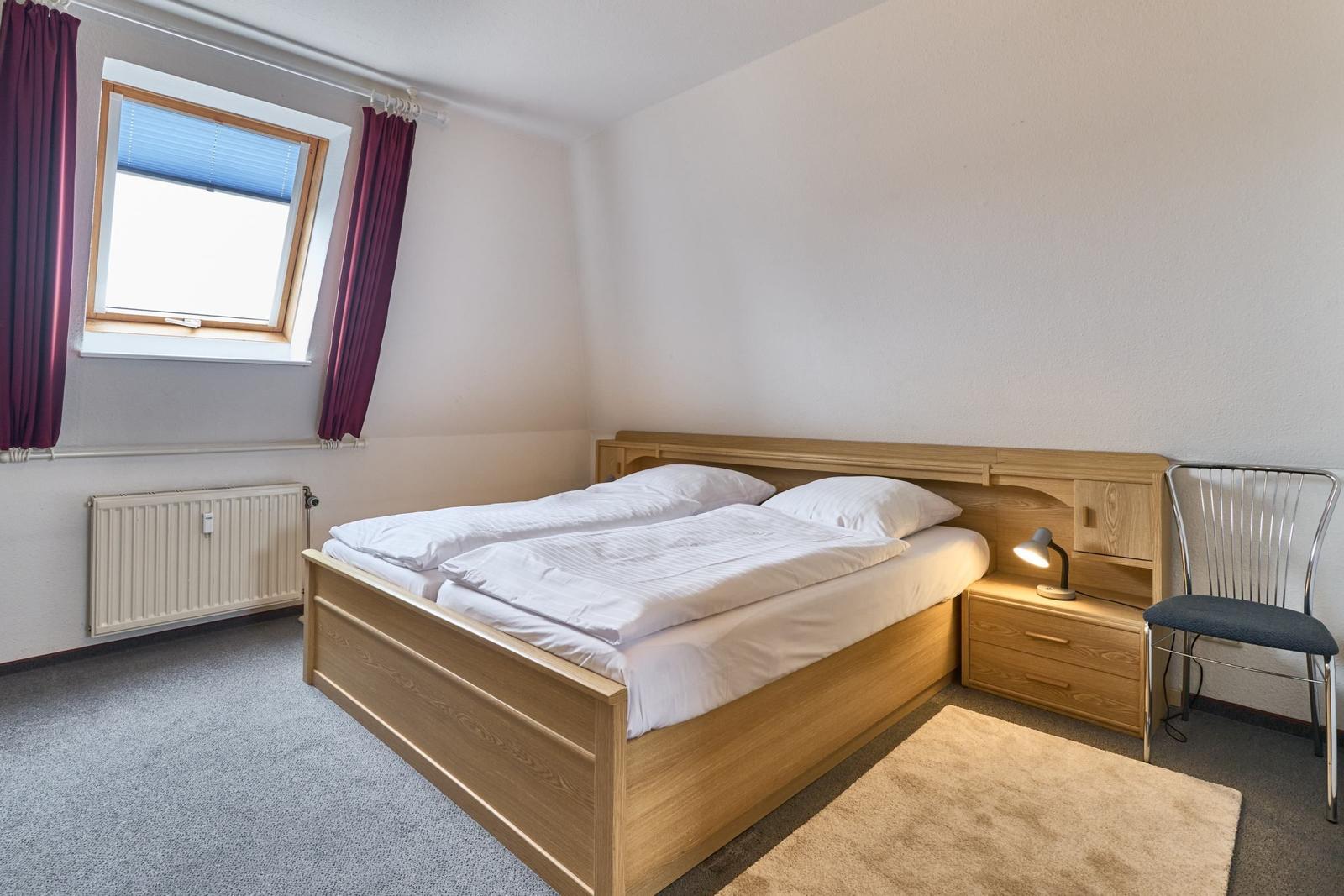 Ein Schlafzimmer mit Doppelbett, Nachttisch und Stuhl. Fenster mit Vorhängen und Heizkörper.