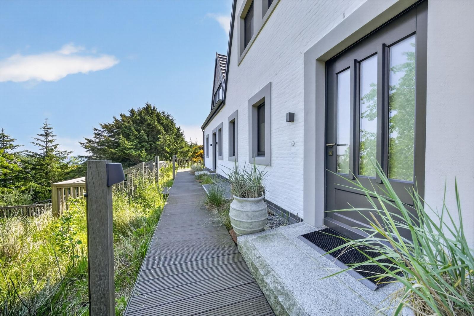 Holzweg führt zur Eingangstür eines weißen Hauses mit grauem Fensterrahmen und Pflanzen.