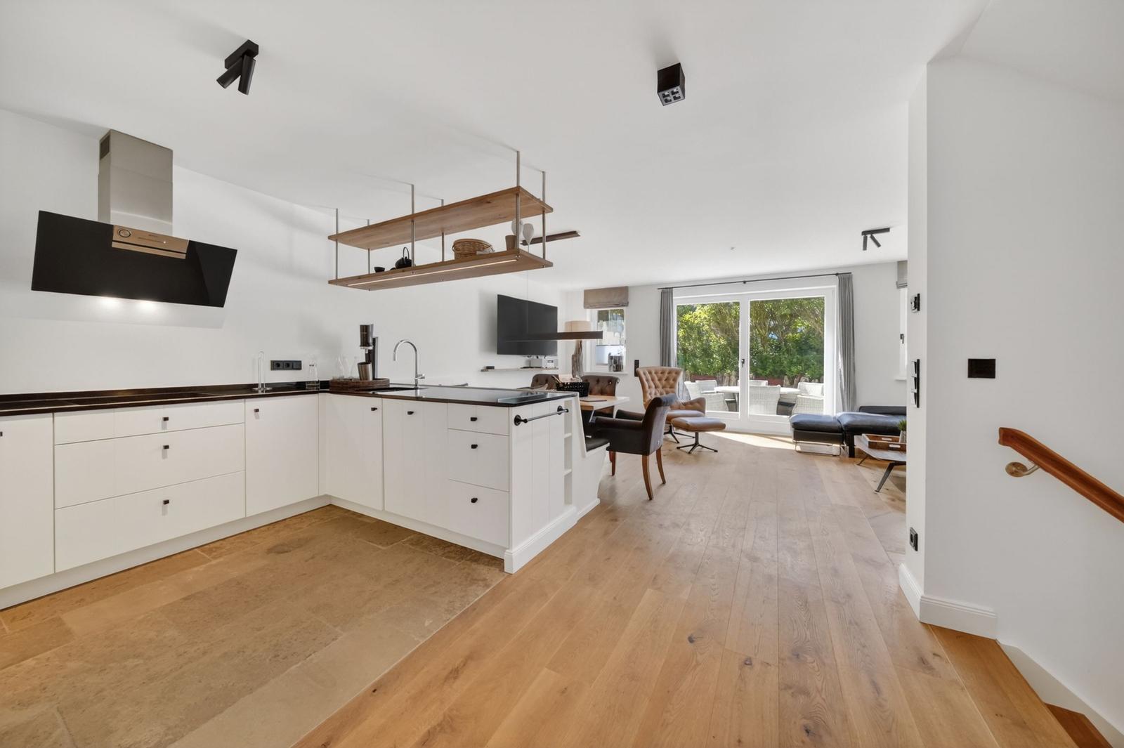 Moderne Küche mit weißen Schränken und Holzboden. Offener Wohnbereich mit Sitzgelegenheiten und Blick auf den Garten.