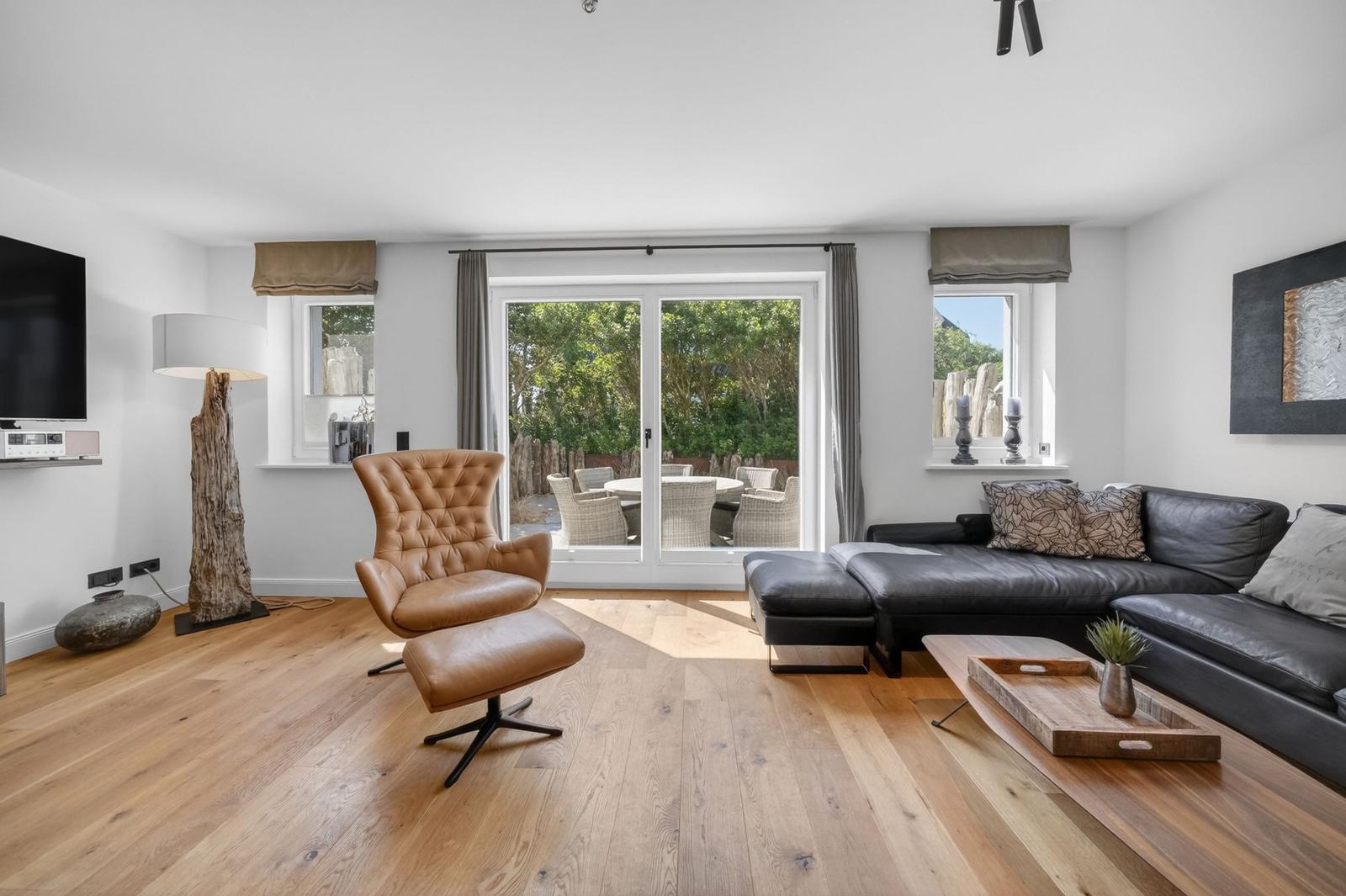 Wohnzimmer mit braunem Sessel, schwarzem Sofa und Holzboden. Durchgang zur Terrasse mit Gartenmöbeln.
