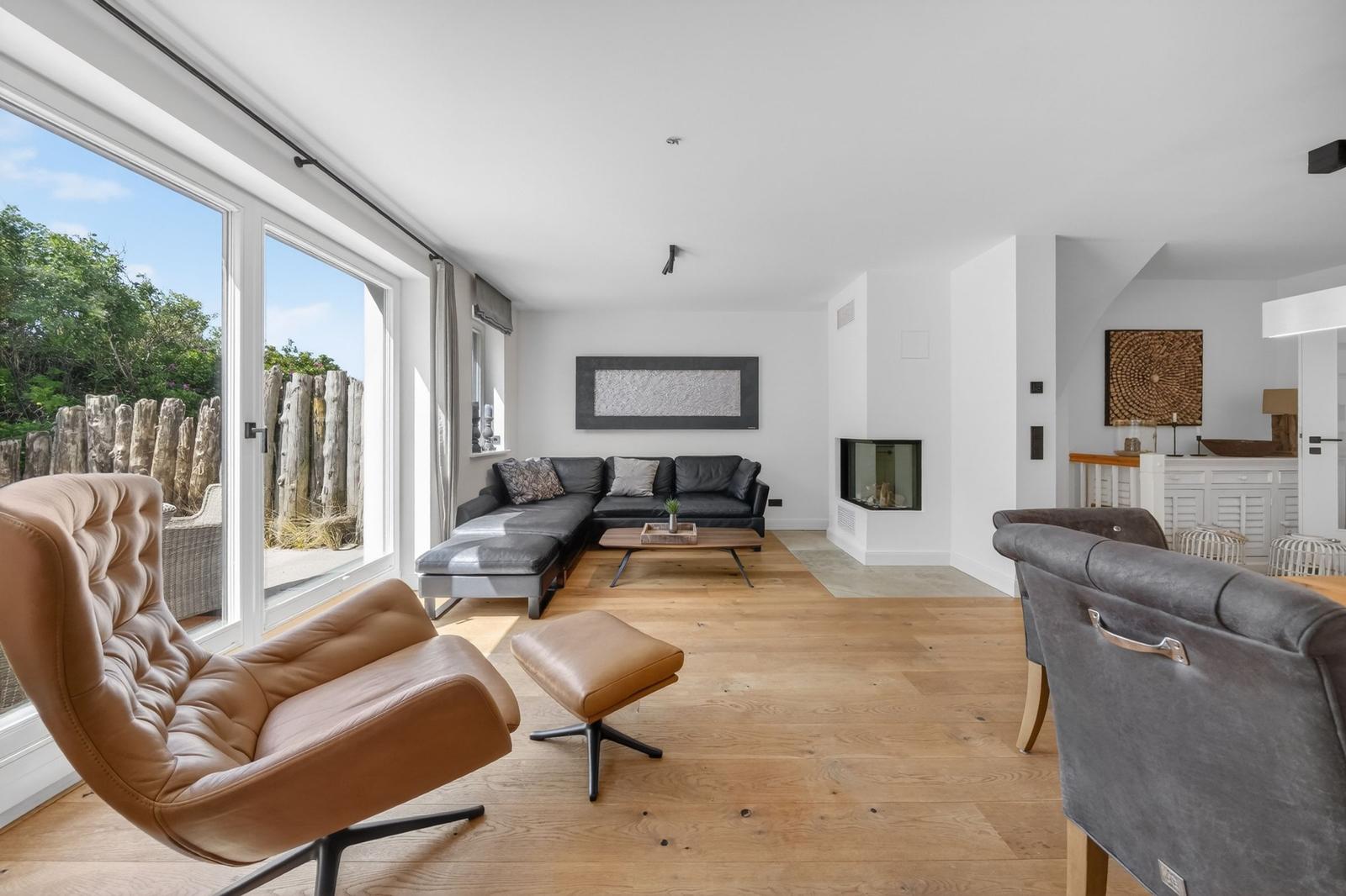 Wohnzimmer mit braunem Ledersessel, schwarzer Couch und Holzboden. Große Fenster und Kamin.