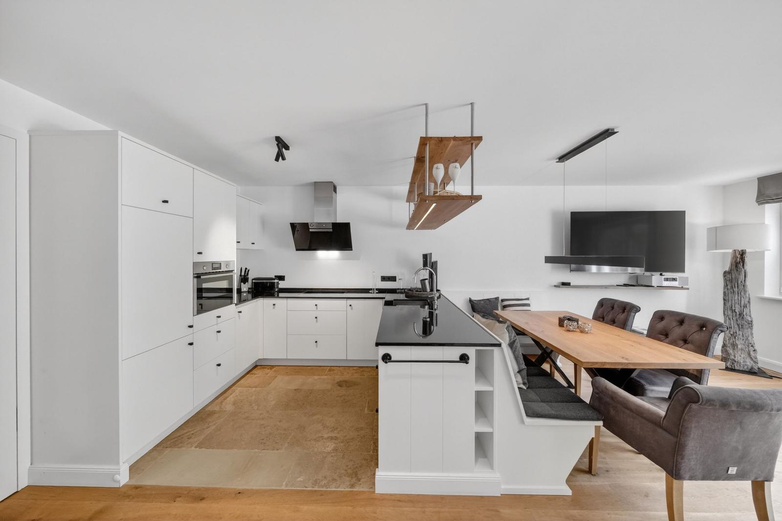 Moderne Küche mit Essbereich und TV. Weiße Schränke, schwarze Arbeitsfläche und Holzstühle.