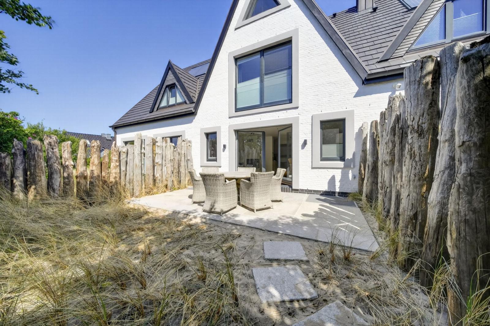 Moderne weiße Villa mit Terrasse und Gartenzaun. Holzstämme umgeben den Außenbereich.
