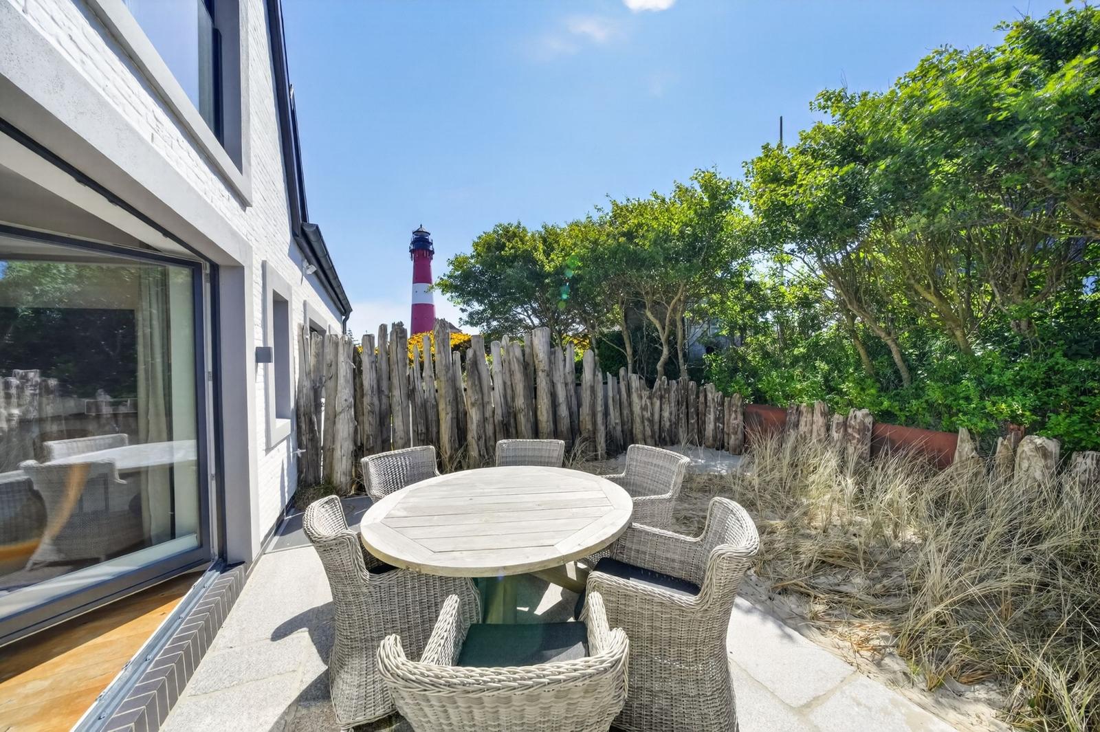 Terrasse mit Tisch und Stühlen, Blick auf Leuchtturm und Grünflächen.
