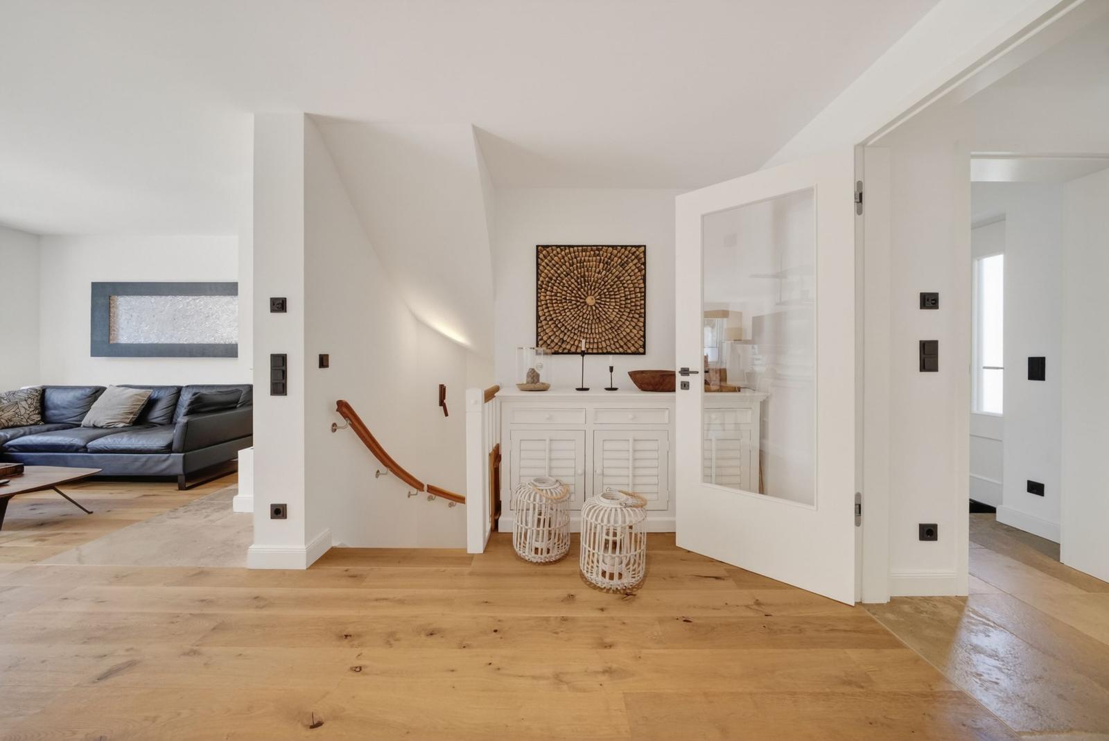 Haus mit Holzboden, weißen Wänden und Treppenhaus. Im Hintergrund ein Wohnzimmer mit Sofa.