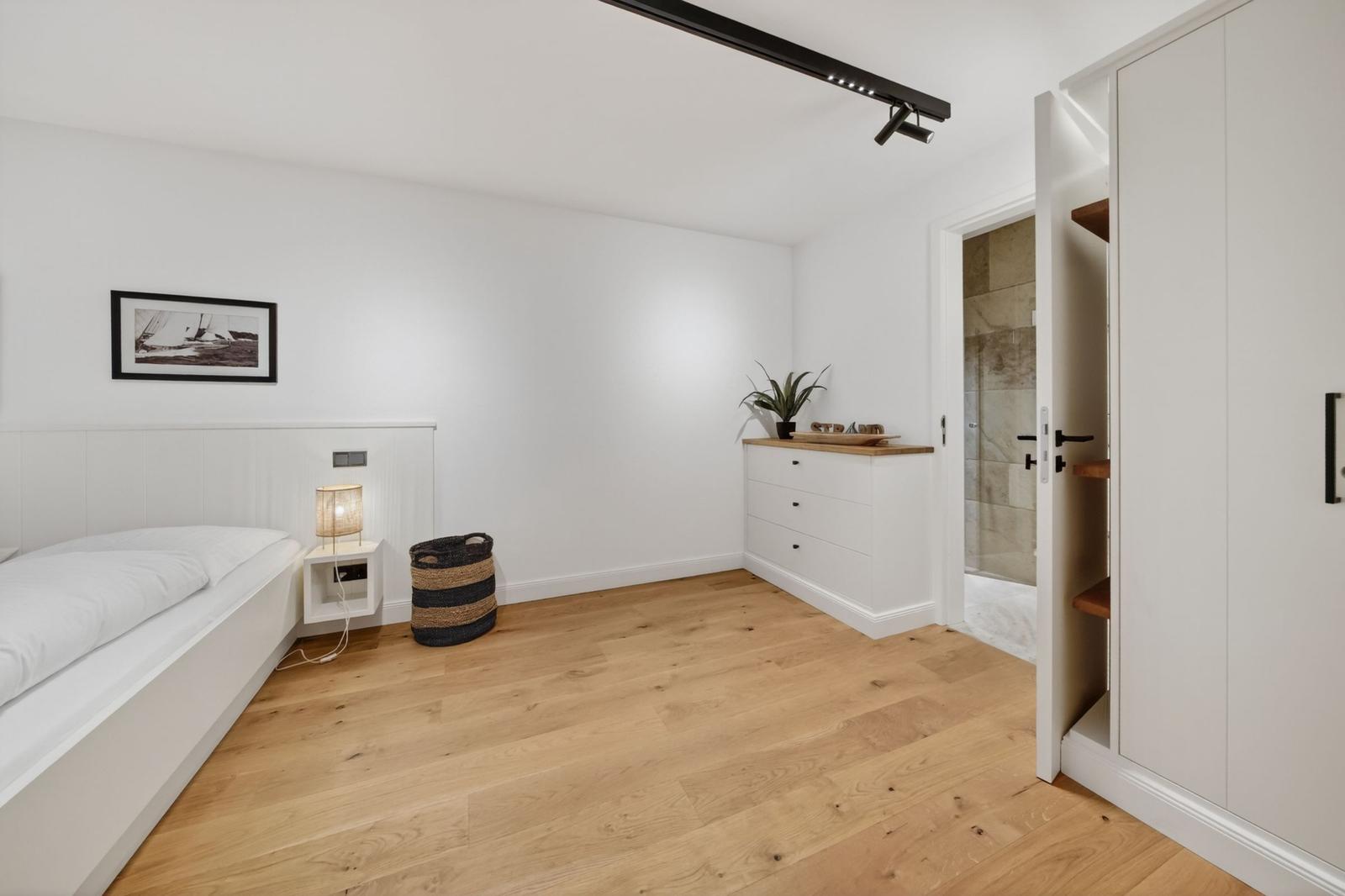 Schlafzimmer mit weißem Bett, Holzboden und Kleiderschrank. Tür führt in Bad.