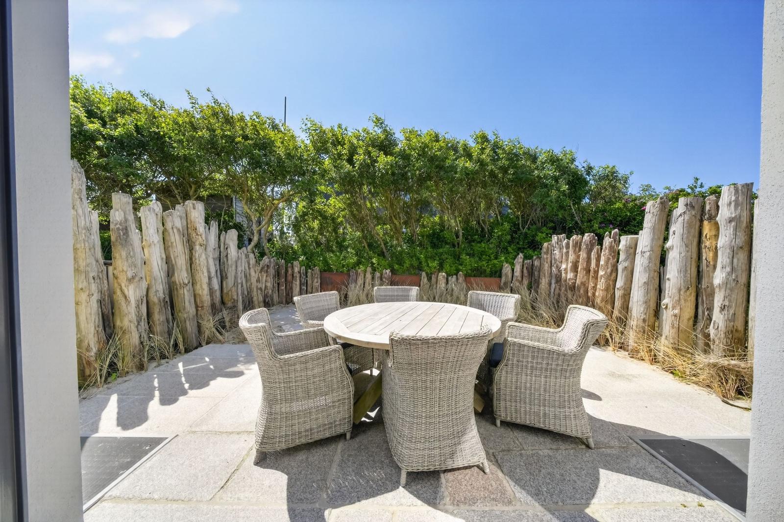Terrasse mit runden Tisch und sechs Stühlen, umgeben von Holzpfosten und Grün.